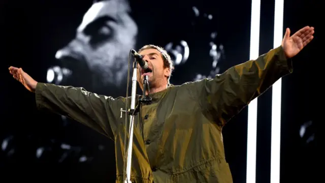 Liam Gallagher in a khaki green raincoat with his arms outstretched