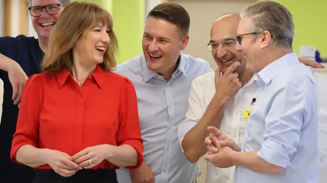 Reeves, wearing red, and Streeting and Starmer, wearing blue shirts, laugh as they walk around a hospital and wellbeing centre in east London