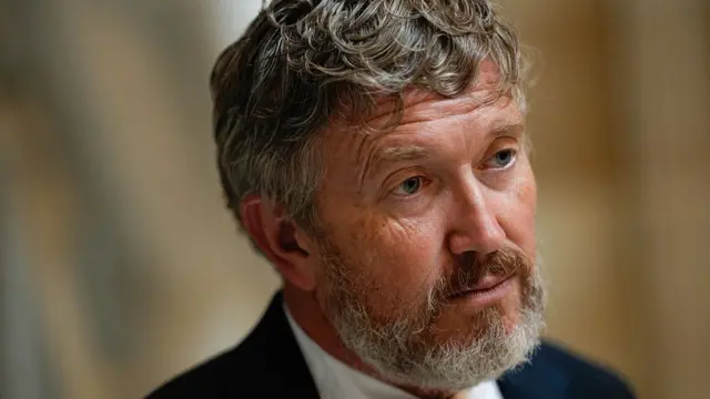 Thomas Massie, wearing a dark suit and white shirt, stares forward.