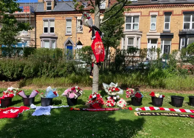 A line of black buckets have been lain out on a grassy verge where fans have placed bunches of flowers. Football shirts and scarves are lain on the grass, and a t-shirt is tied to a small tree in the centre of the frame