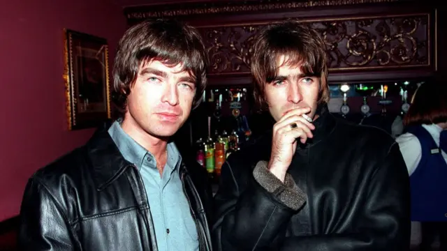 Oasis lead singer Liam Gallagher and brother Noel Gallagher in 1995. They both stand in front of bar full of bottles of alcohol. Noel is wearing a black leather jacket with a blue shirt while Liam is wearing a black leather jacket with fur on the sleeves