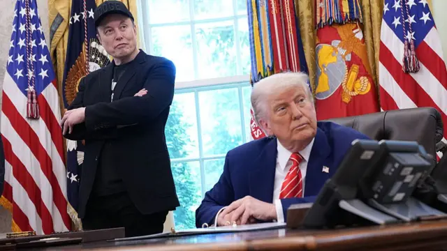Donald Trump sits at a desk while Elon Musk stands next to him