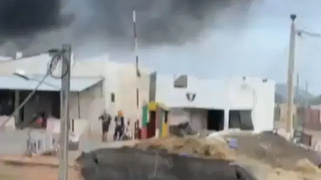 Smoke billows over a series of structures and armed men stand in front of them