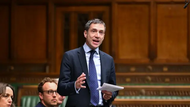 A man wearing a suit speaking, gesturing with one hand and another holding a piece of paper. There's fancy green benches and wooden panelled walls behind him, and a few people are sat behind him