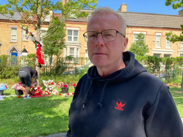 John Barlow, who has light grey hair and is wearing glasses and a dark blue Adidas branded hoodie, looks into the camera with a solemn expression while in the backdrop a man lays flowers by a tree surrounded with tributes.