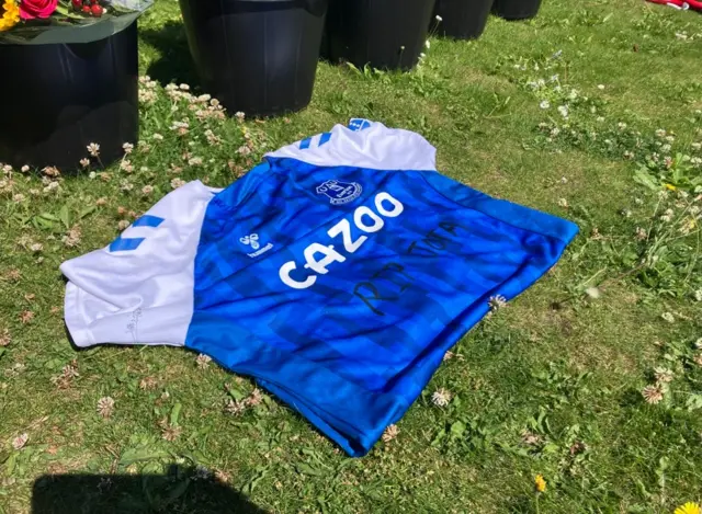 A blue replica Everton kit lying on the grass in front of some black buckets containing bunches of flowers. 'RIP Jota' has been handwritten on the front of the shirt.'