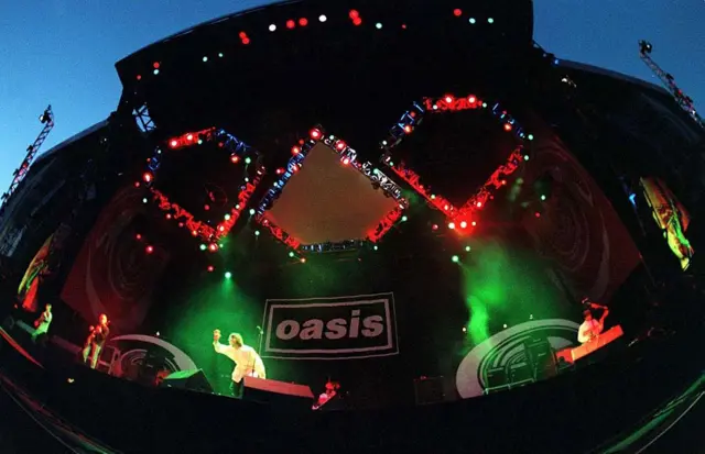 Oasis, performing at Knebworth House, Hertfordshire. The band are on stage with green, red and blue lights behind them. The oasis logo is behind