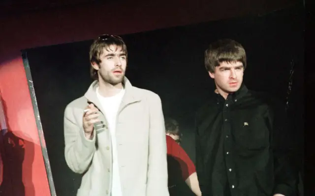 Oasis in 1997. Liam Gallagher is wearing a light coat and white T-Shirt and is holding a cigarette in his right hand, with sun glasses on his head. Noel Gallagher is wearing a black shirt