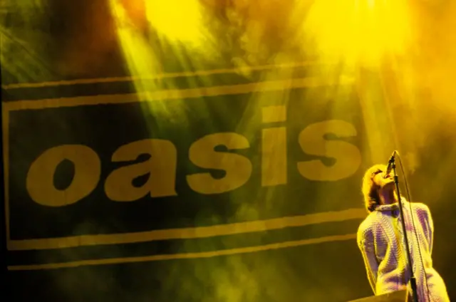 Liam Gallagher performing live onstage, with Oasis logo behind with yellow lights that shine down on him