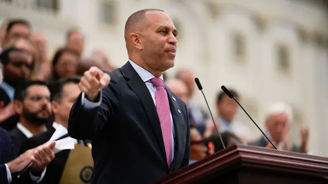 Hakeem Jeffries speaking at a podium wearing a dark suit and a pink tie