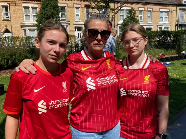 Andrea Molyneux. who is wearing large sunglasses, stands with her arms around the shoulders of her daughters either side of her. All three women are wearing Liverpool kits and have sombre expressions.