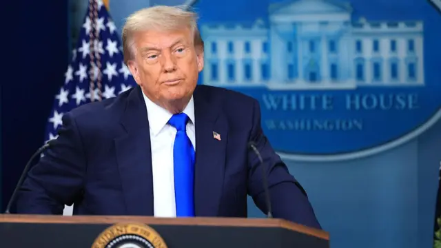 Donald Trump stands at a podium wearing a navy suit and blue tie