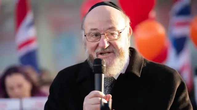 Sir Ephraim Mirvis is seen in closeup, holding a microphone and wearing a kippah