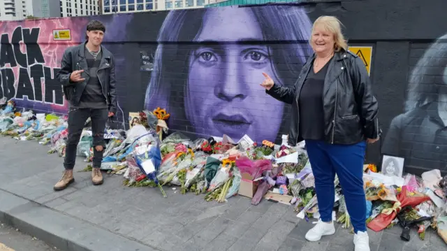 A man in all black with black hair and a woman with blonde hair are standing in front of a purple and black mural of Ozzy Osbourne. There are hundreds of bunches of flowers and photos lying on the ground underneath it. They both have one arm up with the devil horn hand sign in the air.