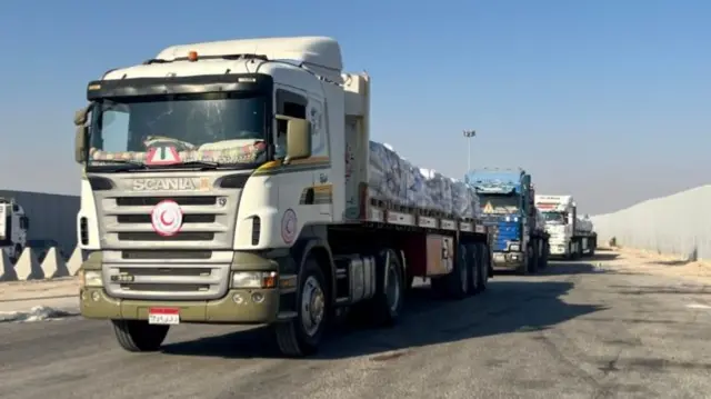 A large white and khaki green truck at the front of a long queue of vehicles with crates of aid in their beds