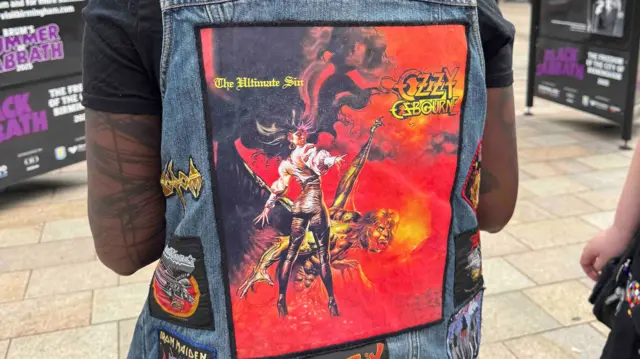 The back of a denim jacket with a large square picture on the back. It is red, orange and yellow. It says "ozzy osbourne" and depicts ozzy as a bat, fangs exposed. There is a woman standing above him with long flowing black hair, her arm outstretched