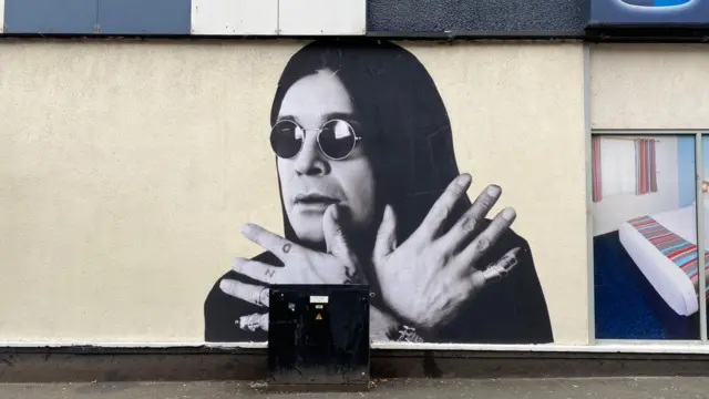 A light yellow wall with a large black and white mural of Ozzy Osbourne on it. He has long black hair and dark, circular glasses on. He is holding both hands out with his fingers splayed.