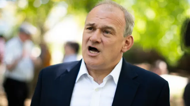 Ed Davey speaking to reporters; he's standing in front of trees and wearing a suit but no tie.