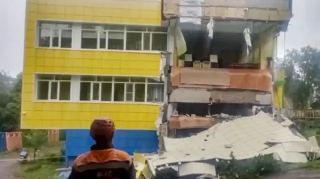 A man stands in front of a destroyed building in Russia.