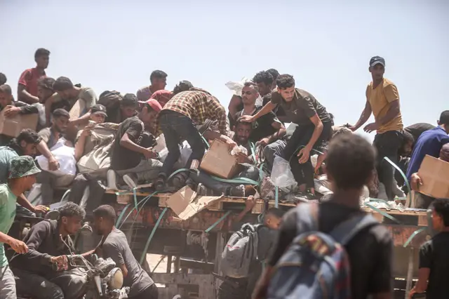 Palestinians gather at an aid distribution point in Gaza's Netzarim Corridor to access a limited supply of flour. There are several men, stepping over one another, as they mad dashedly grab for boxes and bags. The men look distressed. g