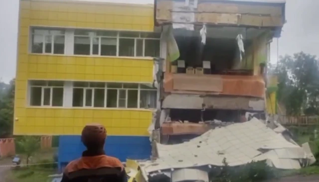 A man in a hard hat looks at a yellow building. The front of the right hand side of the building has come away, meaning that the interior is visible.