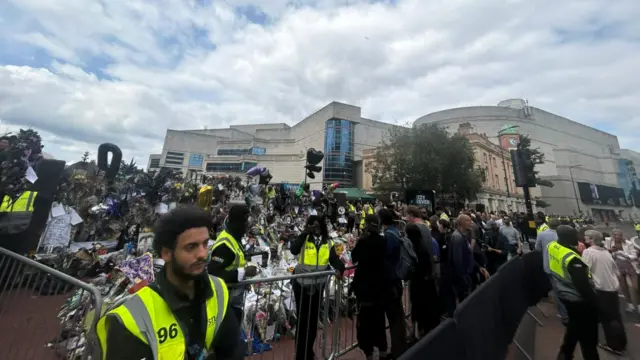 Security guards in hi-viz surround a penned-off area containing floral and other tributes as members of the crowd add to the stack of flowers and memorabilia.
