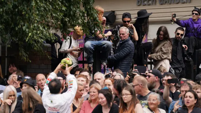 Dozens of people are standing in a crowd, while several above them sit on a black metal fence