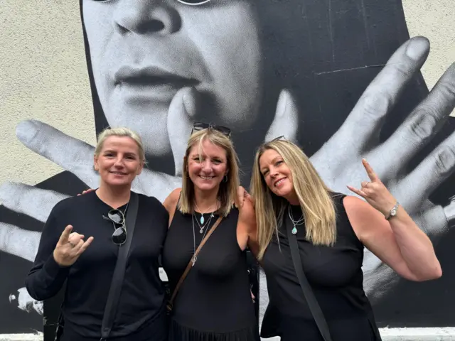 Alex, Jo and Mel, three blonde women with their arms round each other in front of an Ozzy Osbourne mural. Alex and Mel are holding up their index and little fingers in a "rock salute. All three are wearing black tops."