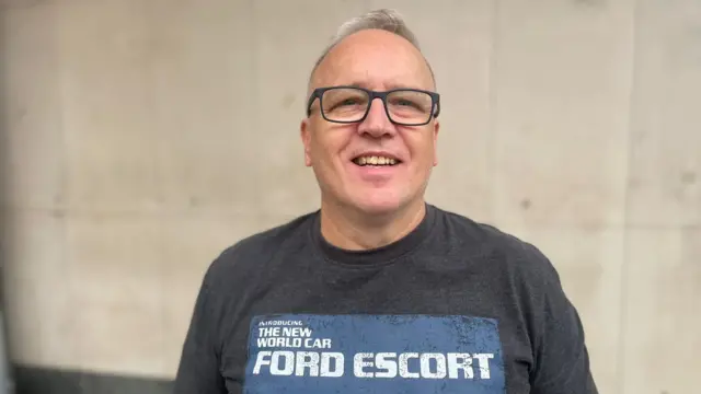 A man with grey hair and black glasses is smiling at the camera. He has a black t-shirt on that reads "the new world car ford escort"