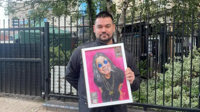 A man with dark hair and facial hair is holding a piece of art. The portrait has a pink background and an image of Ozzy Osbourne on it. He is wearing blue circular sunglasses, all black clothing, gold necklaces and has a cigarette dangling from his mouth