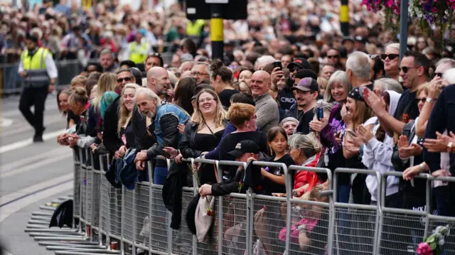 Hundreds of people are standing behind a silver metal fence on a pavement. They are laughing and smiling