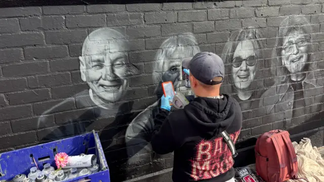 A man with a navy cap is kneeling down in front of a black brick wall, spray painting details onto a black and white mural of Black Sabbath