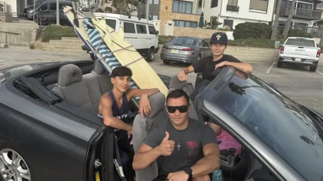 Three males in convertible give thumbs up signs.  They have three surfboards on the back seat.