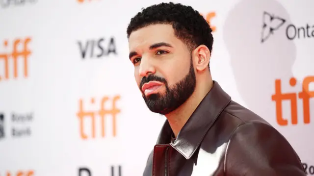 Drake has dark curly hair and dark facial hair and is wearing a maroon leather jacket