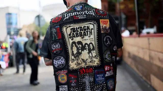 The back of a man who is wearing a black leather jacket. It is covered in patches depicting different heavy metal bands. In the centre is a large picture of Black Sabbath