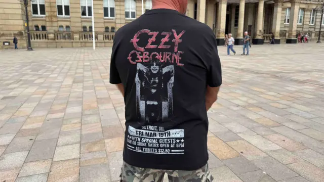 The back of a man wearing a black t-shirt that says "ozzy osbourne" in red writing. There is a black and white picture of ozzy leaning forward, bearing his teeth