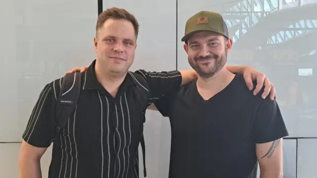 James Hedges, a man in a striped shirt, and John Carr, a man in a black T-shirt and green baseball cap.