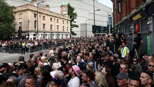thousands of people stand behind metal barriers on either side of a street