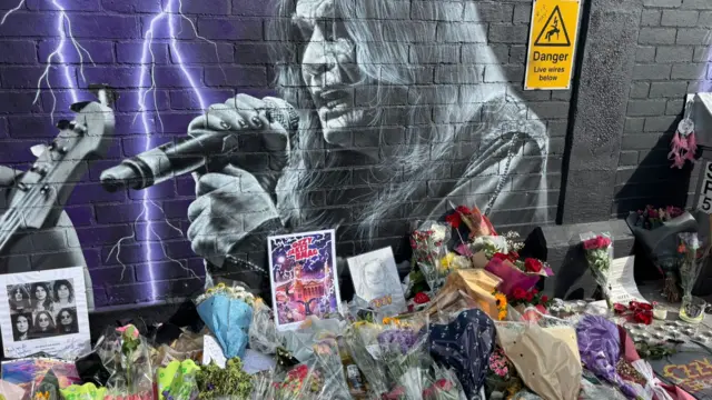 Flowers, cards and photos of Ozzy Osbourne are stacked on the ground underneath a black painted brick wall which has a black and white mural of Ozzy Osbourne singing on it