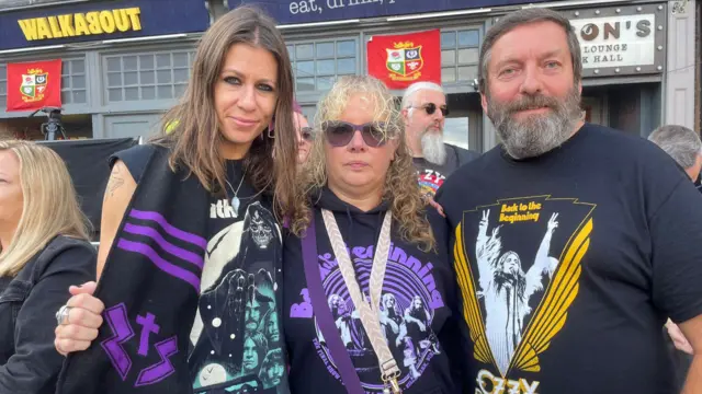 Three people wearing black and purple and black and yellow black sabbath t-shirts. On the left is a woman with long brown hair, in the middle is a woman with curly brown hair and on the right is a man with grey/brown hair and facial hair