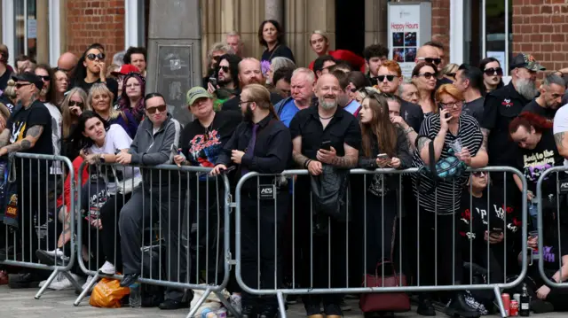 Hundreds of people are standing behind a silver metal fence on a pavement. They are laughing and smiling