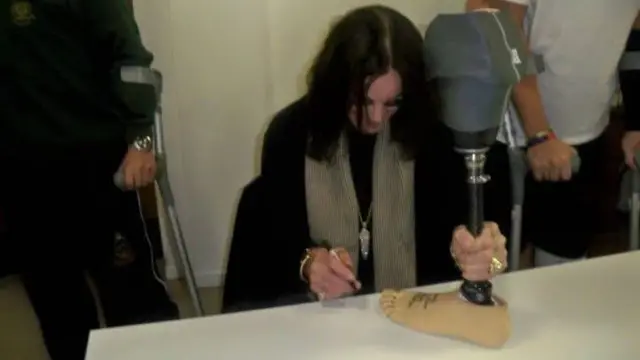 Ozzy Osbourne, who has long black hair, is clutching a black prosthetic leg with a skin-coloured foot on it. He is holding a pen and has signed the foot