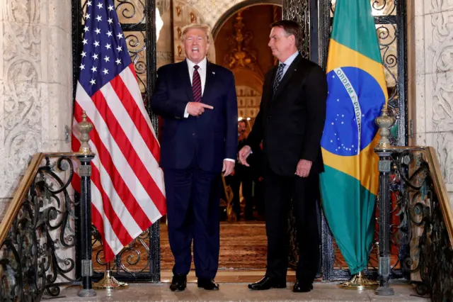 Trump pointing to Bolsonaro on his left, in front of a US and Brazil flag