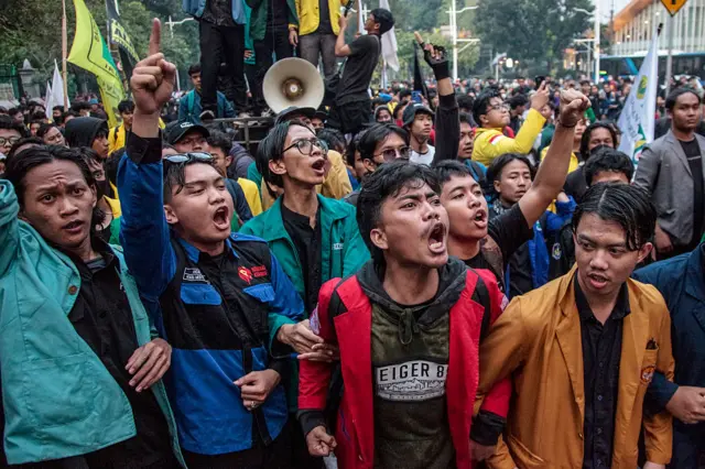 Tight crowd of students in green, blue, red and orange jackets linking arms and opening mouths in shouts