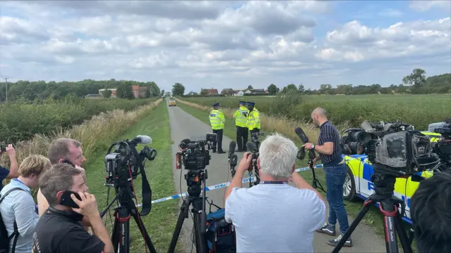 Cameras and microphones set up in front of the police cordon. Three officers are stood beyond it on a single-track lane