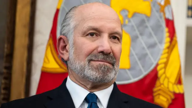 Howard Lutnick stands in front of a flag. He is wearing a black suit over a white shirt and a blue tie.