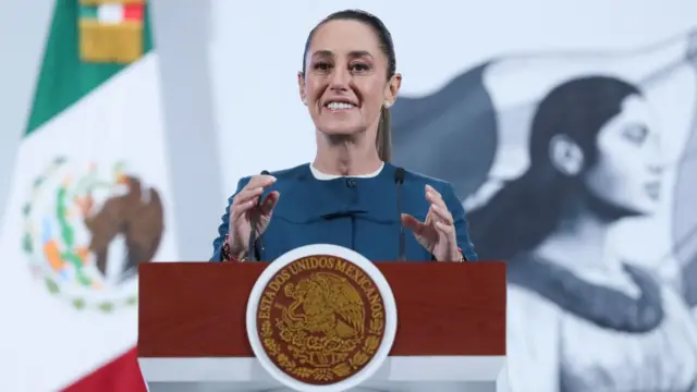 Claudia Sheinbaum smiles while speaking at a podium with a Mexican flag behind her
