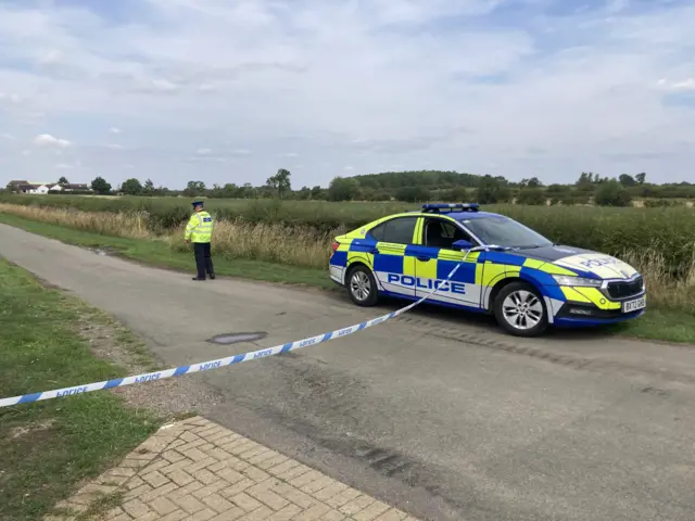 A police tape across a single-track country road. There is also a patrol car parked on the road and one officer.