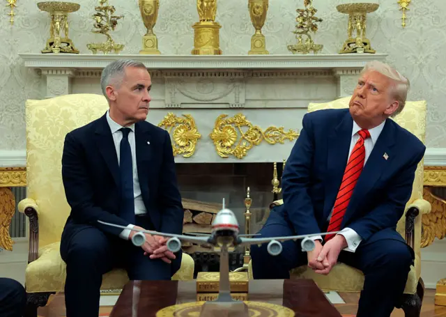 Carney in dark suit and tie, on gold chair and in front of Oval Office's golden decor, looks over at Trump, in a blue suit and red tie, who is leaning away and frowning. A plane model is between them