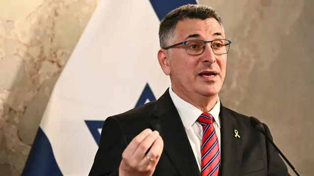 Israel's Foreign Minister Gideon Sa'ar speaking into a small microphone with an Israeli flag hung behind him.